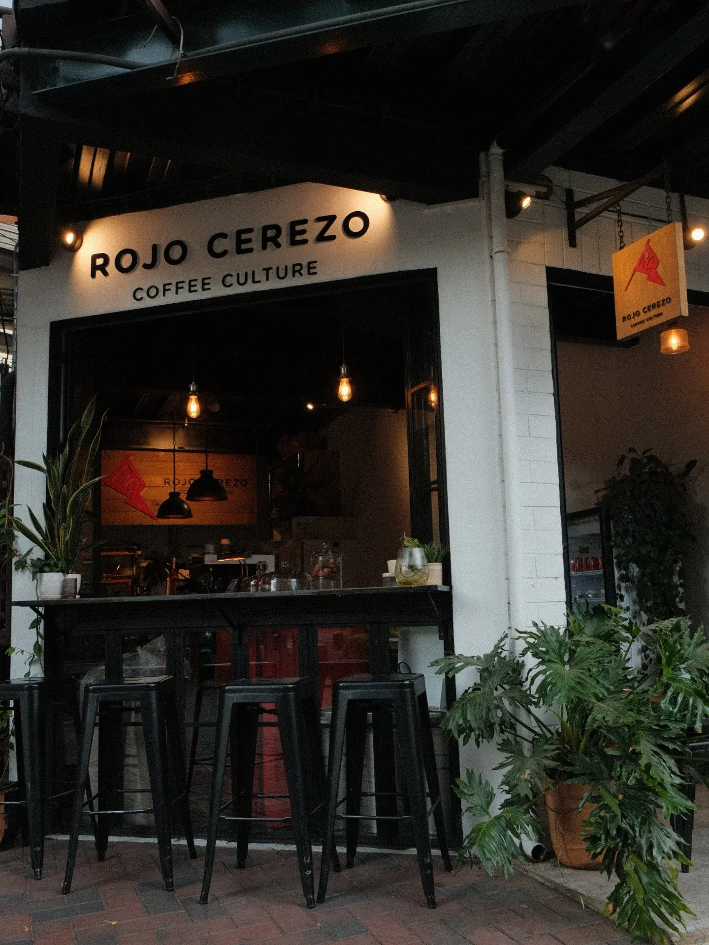 Fachada de Rojo Cerezo Coffee: rótulo Coffee Culture, ventanal iluminado y bancos rojos en la acera
