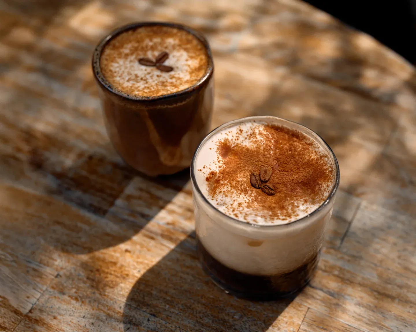 Dos bebidas con espuma y canela sobre una mesa de madera al sol